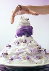 Fioletowy słoik z etykietą "EQUALBERRY DEEP CERA BAKUCHIOL Plumping" stoi na górze białej góry bitej śmietany z fioletowymi kwiatowymi akcentami.