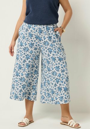 Kobieta w białych, szerokich spodniach o długości 7/8 z niebieskim kwiatowym wzorem, granatowym bezrękawnikiem, złotą bransoletką i białymi sandałami typu slide.