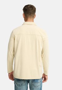 Mand iført en beige kordfløjl skjorte med lange ærmer og blå jeans, står med ryggen til på en ensfarvet lyse baggrund.