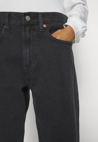 Los jeans de mezclilla negra presentan un diseño de pierna recta, herrajes de cobre y bolsillos laterales. La tela tiene una textura sutil con un acabado clásico.