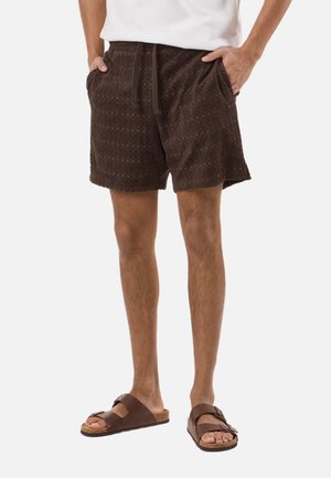Mann trägt dunkelbraune Shorts mit Struktur und Seitentaschen sowie braune Sandalen mit zwei Riemen, steht mit den Händen in den Taschen.