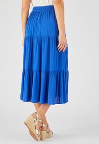 Falda azul en varios niveles con cintura fruncida, hecha de tejido ligero y texturizado. Se lleva con sandalias doradas de cuña con tiras en el tobillo.