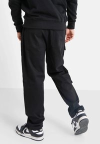 Pantalones cargo negros con cintura elástica, múltiples bolsillos y puños acanalados. Combinados con zapatillas negras y blancas que presentan una marca visible.