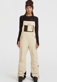 Beige geïsoleerde sneeuwoverall met verstelbare banden, een bruine borstzak, twee zijritsen en oranje accenten. Draag over een zwart shirt met lange mouwen.