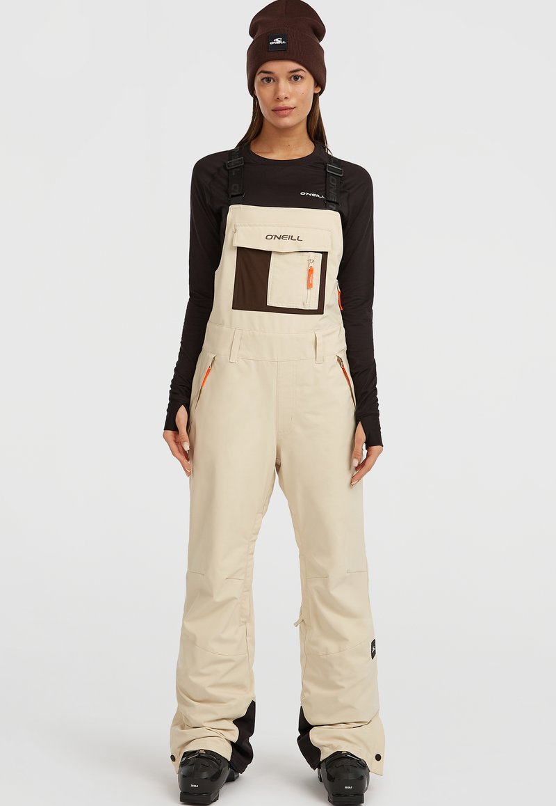 Beige geïsoleerde sneeuwoverall met verstelbare banden, een bruine borstzak, twee zijritsen en oranje accenten. Draag over een zwart shirt met lange mouwen.