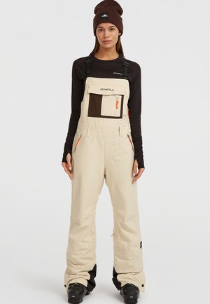 Macacão isolante de neve em bege, com alças ajustáveis, um bolso de peito castanho, dois zíperes laterais e detalhes em laranja. Usado sobre uma camisa preta de mangas longas.