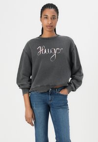 Dark gray sweatshirt with a crew neck and oversized sleeves, featuring a cursive "Hugo" logo in light pink. Paired with blue denim jeans.