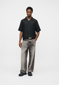 Άνδρας που στέκεται με το ένα χέρι στην τσέπη, φορώντας μαύρο πουκάμισο με κοντά μανίκια και κουμπιά, ξεθωριασμένο παντελόνι με φαρδιά μπατζάκια, μαύρα παπούτσια και ζώνη με ασημένια αγκράφα.