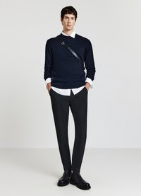 Marineblauer Pullover mit V-Ausschnitt, kombiniert mit gemusterten schwarzen Hosen und schwarzen Lederstiefeln. Verfügt über einen horizontalen Schultergurt.