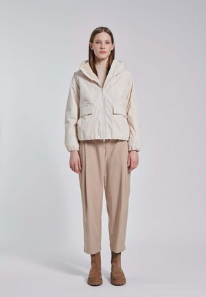 Donna in piedi che indossa una giacca con cappuccio color crema, pantaloni beige e stivali in camoscio marrone contro uno sfondo chiaro e semplice.
