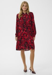 Rochie roșie cu flori, cu mâneci lungi, guler cu nasturi și croială lejeră. Prezintă flori mari, alb-negru, pe un material texturat.
