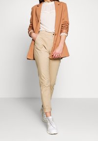 Beige chinos met een tapered fit, gecombineerd met een licht textuurblouse en een camel blazer. Geaccentueerd met witte sneakers.