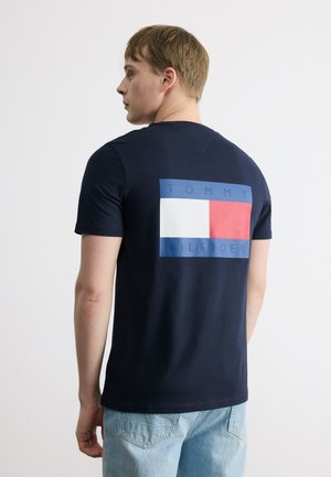 Młody mężczyzna stojący bokiem, ubrany w granatowy t-shirt Tommy Hilfiger z dużym logo w kolorach czerwonym, białym i niebieskim na plecach, w połączeniu z jasnoniebieskimi dżinsami.