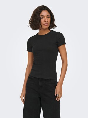 Vrouw met kort krullend haar, gekleed in een nauwsluitend zwart T-shirt met korte mouwen en zwarte broek, kijkt naar rechts met een neutrale uitdrukking.