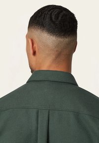 Donkergroene, textuurrijke shirt met een kraag, rugplooien en zichtbare stiksels. Het nauw geschoren haar van het model is zichtbaar.