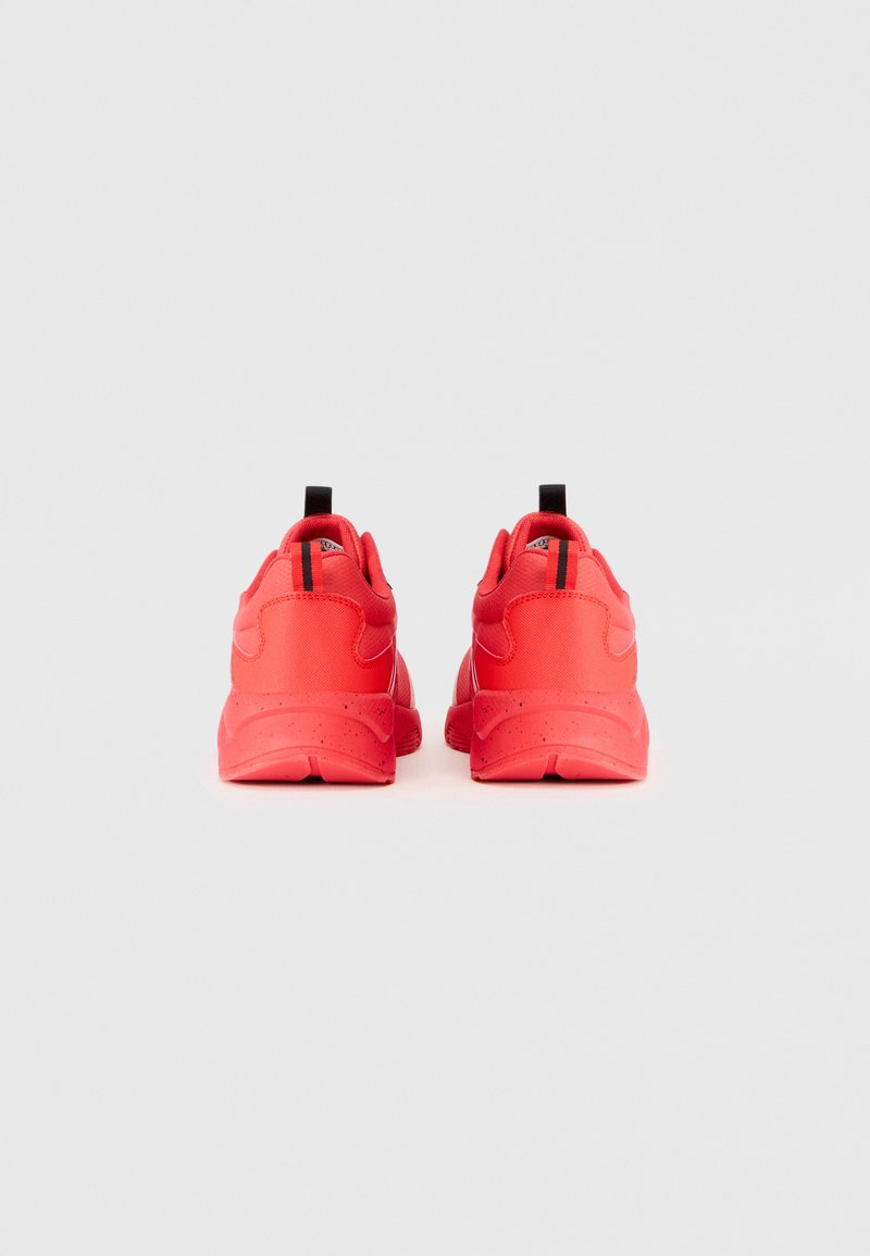 Des chaussures de sport rouges avec une surface texturée, un dos arrondi et une poignée noire. Elles possèdent un col rembourré et une bande noire contrastante sur chaque talon.