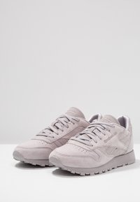 Ljusgrå mockaskor med en texturerad ovandel, rund tå, snörning och gummisula med greppmönster. Reebok-logotyp synlig.