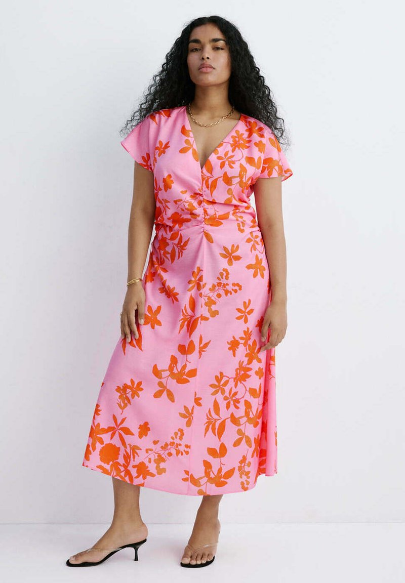 Femme aux longs cheveux bouclés portant une robe midi rose à imprimé floral orange et des talons noirs ouverts à l’avant, debout sur un fond blanc.