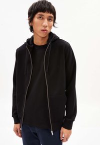 Felpa nera con zip, dotata di tasca frontale, cappuccio con cordino e texture liscia, indossata sopra una maglietta nera, abbinata a pantaloni blu navy.