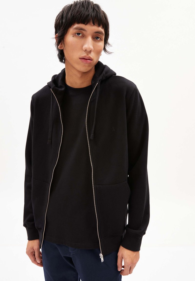 Felpa nera con zip, dotata di tasca frontale, cappuccio con cordino e texture liscia, indossata sopra una maglietta nera, abbinata a pantaloni blu navy.