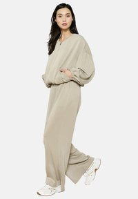 Tutina beige con una vestibilità comoda, spalle scese e polsini elasticizzati. Include tasche laterali e un dettaglio con zip davanti. Indossata con sneakers bianche.