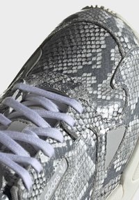 Grå och vit sneakers med texturerat ormskalemönster på ovansidan, vita snören och en reflekterande Adidas-logotyp på sidan.