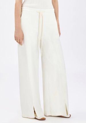 Pantaloni bianchi a gamba larga con spacchi frontali, tasche laterali e vita con coulisse, indossati con scarpe color cuoio e un top bianco a coste.