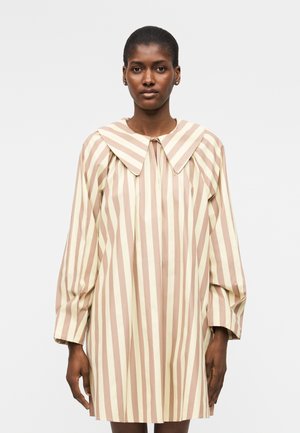 Frau trägt ein lockeres, langärmeliges Kleid mit breiten beigen und cremefarbenen vertikalen Streifen und einem übergroßen spitzen Kragen.