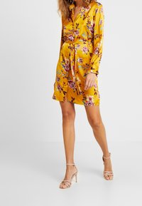 Blommig omlotklänning i livlig gul satin, med långa ärmar, bindning i midjan och knälång nederdel, prydd med ros- och bladmönster.