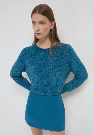 Maglione corto lavorato a maglia color teal con un design texturizzato a coste. Abbinato a una mini gonna teal in tinta unita. Entrambi i capi hanno maniche lunghe.