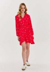 Červené šaty s dlouhými rukávy, květinový vzor, volantový lem, zavázaný pas, vyrobené z lehké tkaniny, nošené s bílými teniskami.