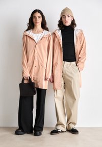 Two individuals wear matching light peach waterproof jackets with hoods, paired with dark trousers and casual footwear. A textured black bag adds detail.
