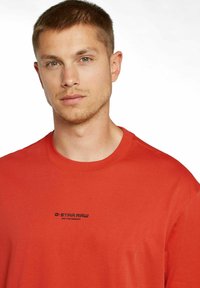 T-shirt rossa a maniche corte in cotone, caratterizzata da un collo rotondo e un logo nero con testo sulla parte anteriore. Texture liscia, design casual.