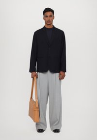 Blazer nero sopra una maglietta scura, pantaloni palazzo grigi chiari e borsa a tracolla in pelle color tan. Il modello è in piedi di fronte a uno sfondo semplice.