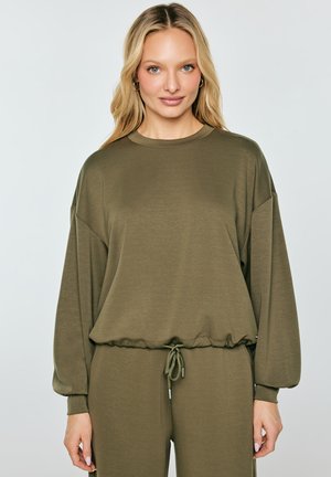 Blondynka w oliwkowozielonej bluzce z długim rękawem, z wiązaniem w talii, i dopasowanych spodniach, stojąca na jasnym, jednolitym tle.