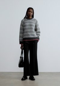 Grå och svart mönstrad tröja med rosa kant, kombinerad med svarta vida byxor och en svart handväska; modellen står mot en neutral bakgrund.