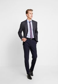 Completo blu navy con un blazer scuro texturizzato, camicia azzurra chiara e cravatta a righe. Cinture nera e scarpe completano il look formale.
