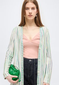 Νεαρή γυναίκα με μακριά μαλλιά που φορά ριγέ ζακέτα, ροζ τοπ, σκούρο τζιν, σκουλαρίκια από μαργαριτάρια, κρατώντας ένα μικρό πράσινο τσάντα με ρυτιδώσεις.