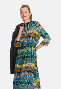 Bunt gemustertes Maxikleid mit Kordelzug in der Taille, in den Farben Blau, Grün, Orange und Schwarz. Das Modell hält einen marineblauen Blazer.