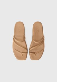 Beige kumikengät, joissa on teksturoitu pohja, ristiin menevät hihnat ja minimalistinen muotoilu. Ei näkyvää metallia tai koristeita.