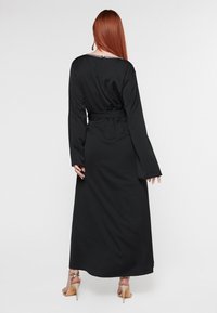 Musta pitkähihainen mekko, jossa on vyötäröllä vyö, sileä pinta ja kaula-aukko, jossa on koristeellisia elementtejä. Elegantti virtaus helmaan.