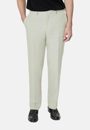 Mężczyzna w jasnobeżowych eleganckich spodniach z zakładkami z przodu, czarnych eleganckich butach i czarnej koszuli, ręce częściowo w kieszeniach.