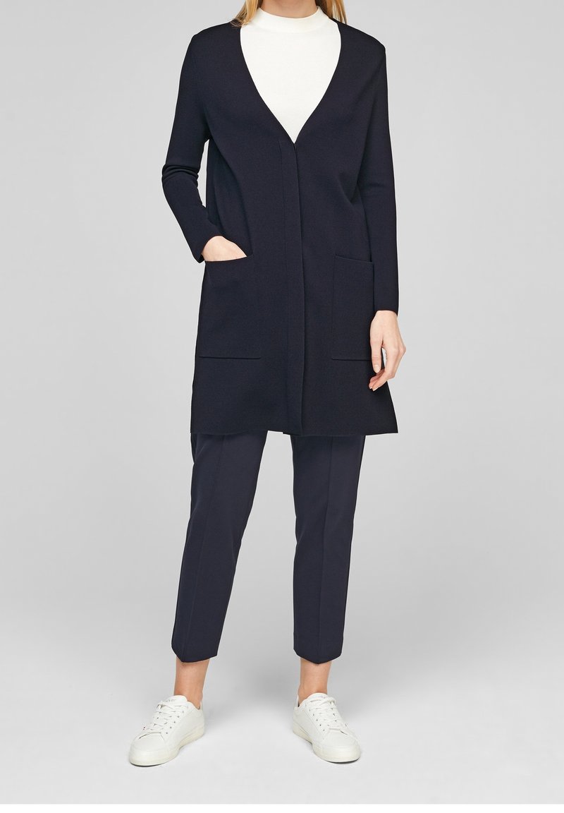 Marinblå lång cardigan med V-ringning, två frampocketar och vanlig passform, kombinerad med mörka byxor och vita sneakers.