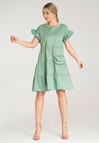 Zielona sukienka na warstwy z bufiastymi rękawami, wykonana z lekkiego materiału. Połączona z małą torebką noszoną na krzyż i odkrytymi beżowymi szpilkami.