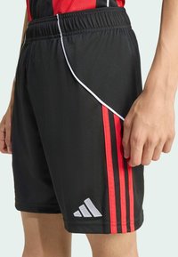 Svarta sportiga shorts med röda sidostripes, elastisk midja och en vit accentlinje. Inkluderar en silver logo patch. Slät tygstruktur.