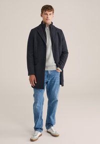 Cappotto di lana grigio scuro con revers, indossato sopra un maglione grigio chiaro, abbinato a jeans blu e scarpe da ginnastica bianche. Texture morbide, forma aderente.