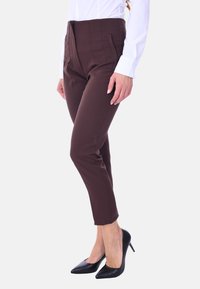 Pantaloni tailored marroni con vita alta, pieghe frontali e tasche laterali, abbinati a tacchi neri a punta e una camicia bianca.