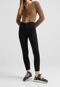 Jeans negros ajustados de cintura alta, combinados con un cárdigan corto y peludo de color marrón. Llevados con zapatillas gruesas en blanco y negro.