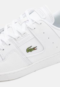 Gros plan d'une basket en cuir blanc avec des lacets blancs et un logo crocodile vert brodé sur le côté.