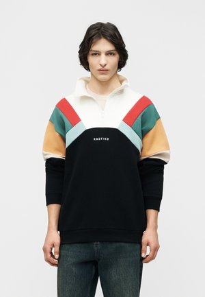 Fiatal férfi sötét hajjal, fekete, fehér, piros, türkiz és bézs színű, színblokkolt cipzáras pulóvert visel, amelyen a "KAOTIKO" felirat olvasható.